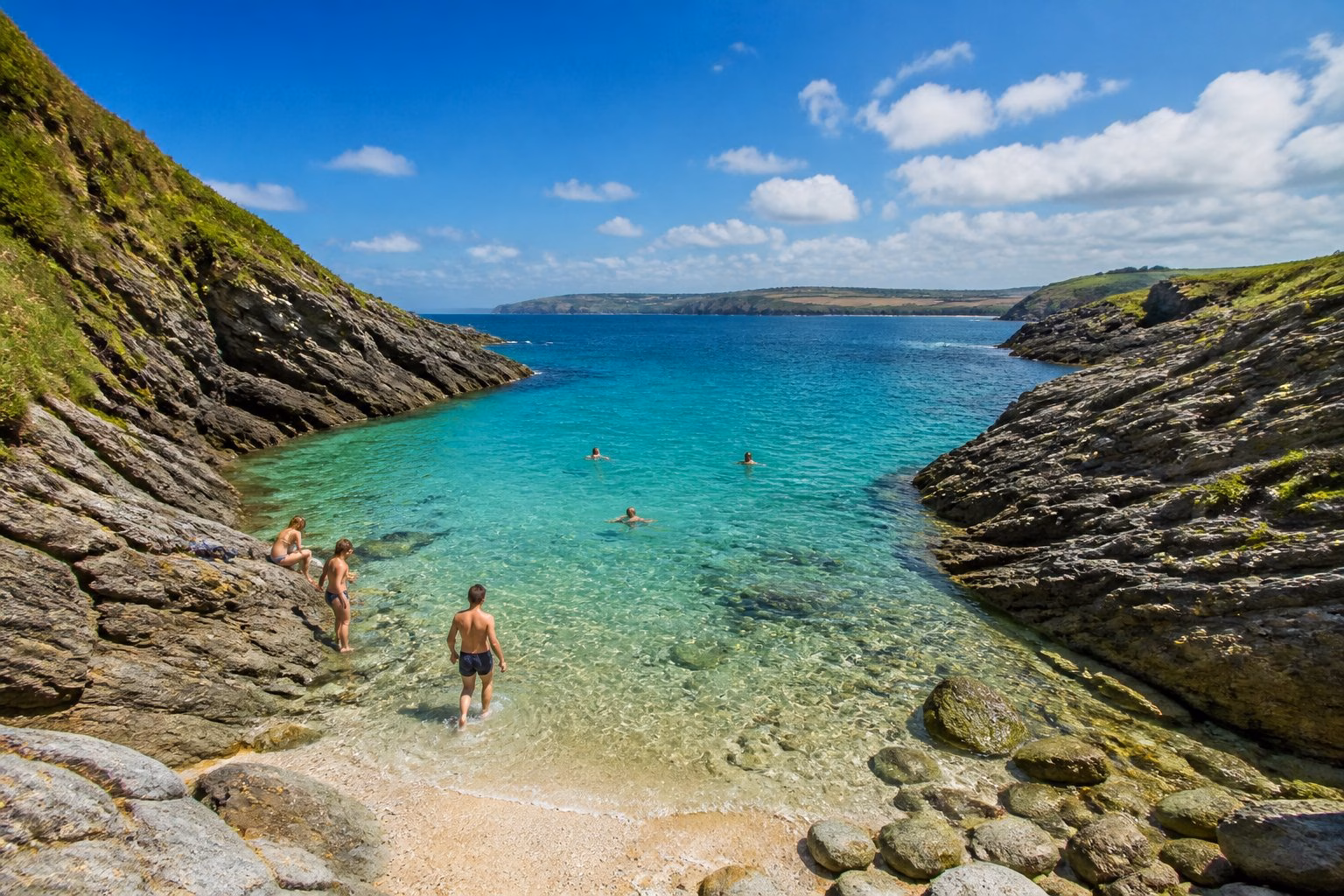 A clear turquoise cove surrounded by rocky cliffs and green vegetation with people swimming and enjoying the water.