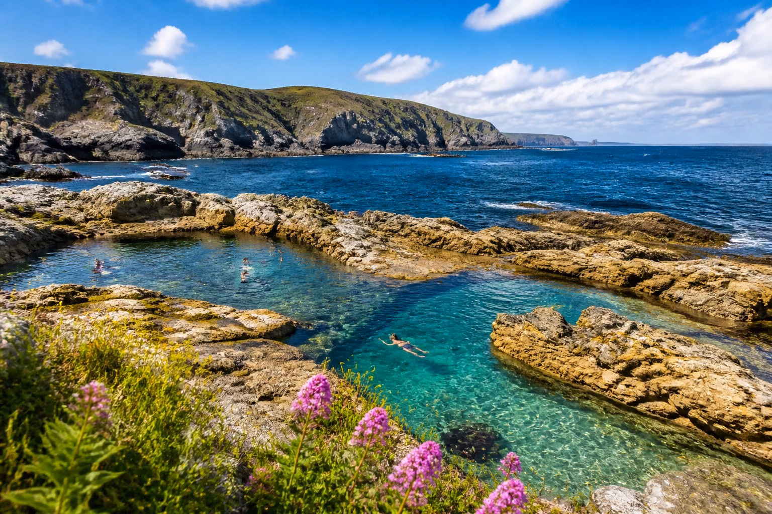 Clear seawater in natural rock pools along the rugged Cornish coastline with cliffs and a bright sky in the background.