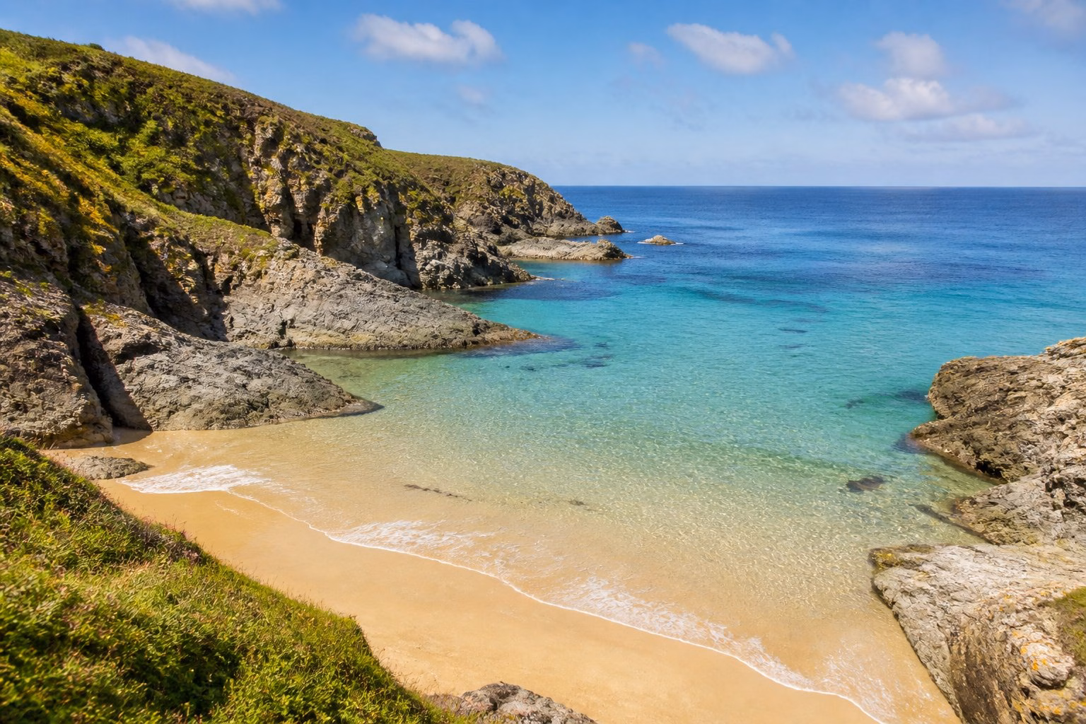 Secluded sandy beach with clear turquoise water, rocky cliffs, and green vegetation under a blue sky.