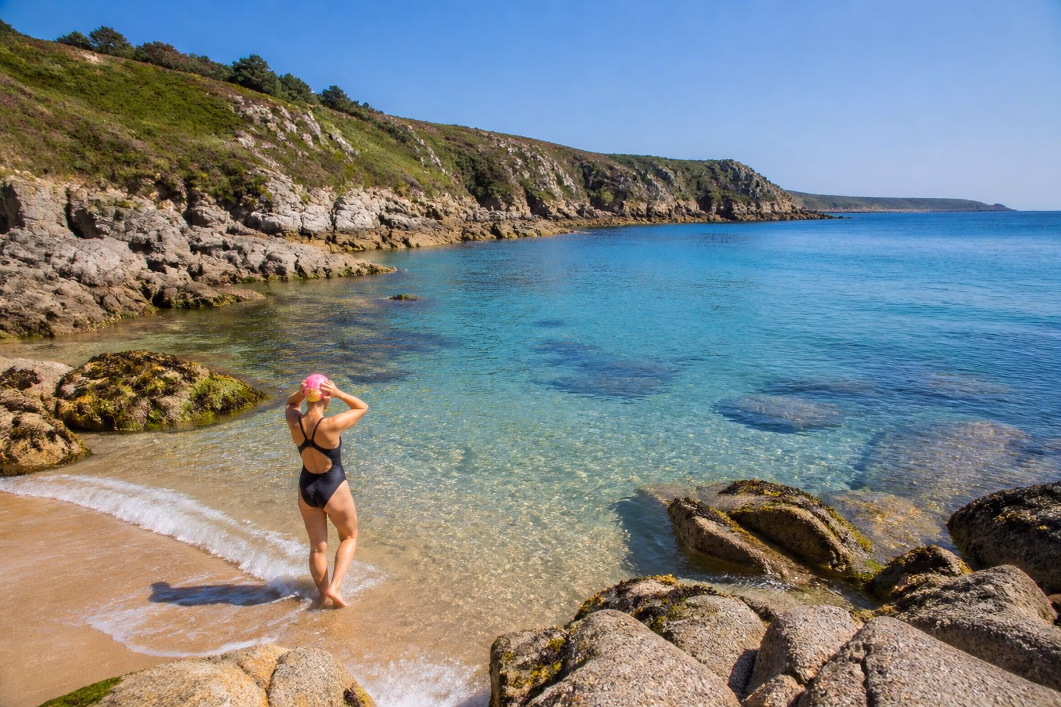 A swimmer preparing to enter clear coastal waters surrounded by rocky cliffs and green vegetation on a sunny day in Cornwall.