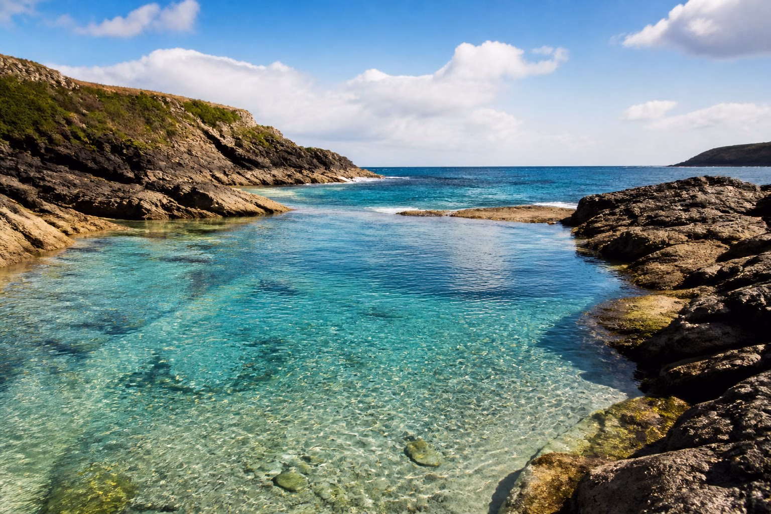 A calm natural swimming spot in Cornwall with clear water, rocky shores, and green plants under a partly cloudy sky.