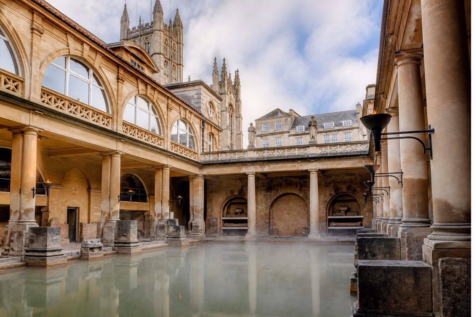 The historic Roman Baths in Bath, UK, showing a large stone thermal pool surrounded by ancient stone architecture and greenery, with no people swimming.