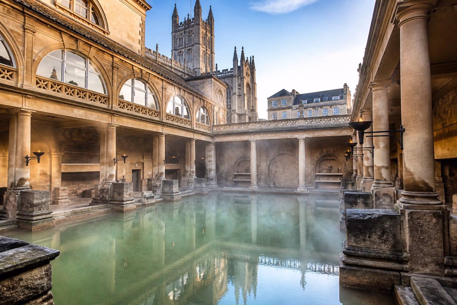 The Roman Baths in Bath, UK, showing the ancient stone pool with steam rising from the warm water surrounded by classical columns and greenery.