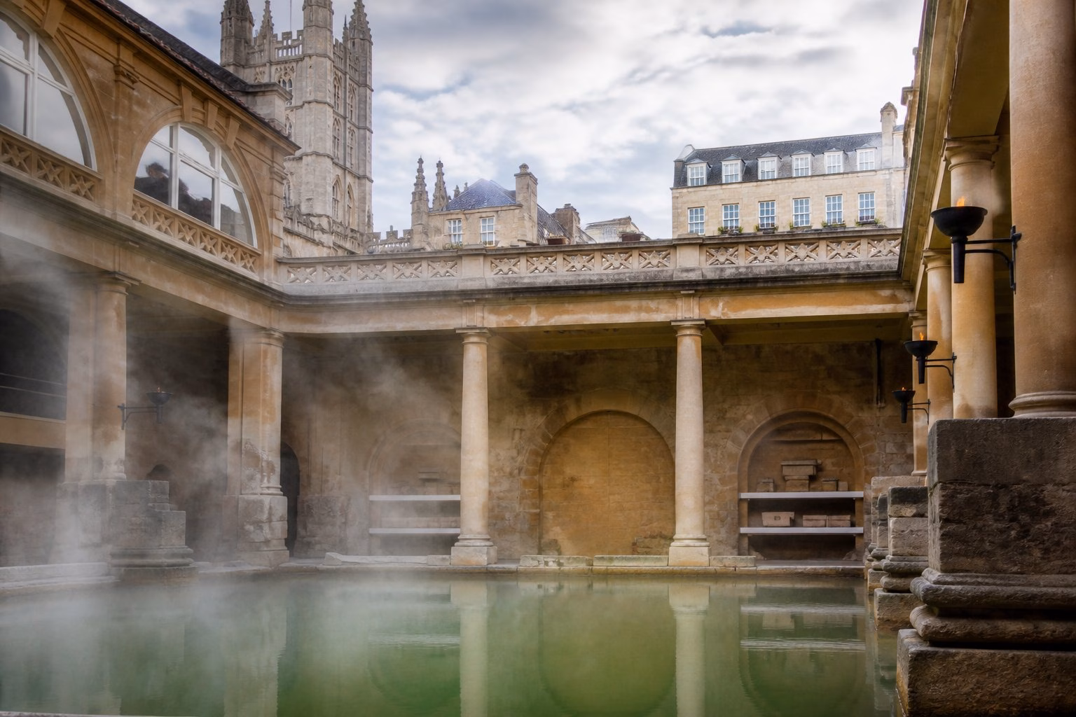 The Roman Baths in Bath, UK, showing a large pool of clear water surrounded by ancient stone architecture and greenery.