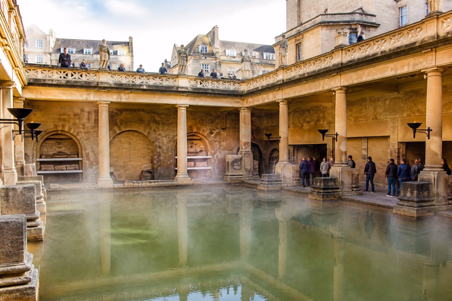 Historic Roman Baths in Bath, UK, showing ancient stone architecture and thermal water pools with visitors walking nearby but not swimming.