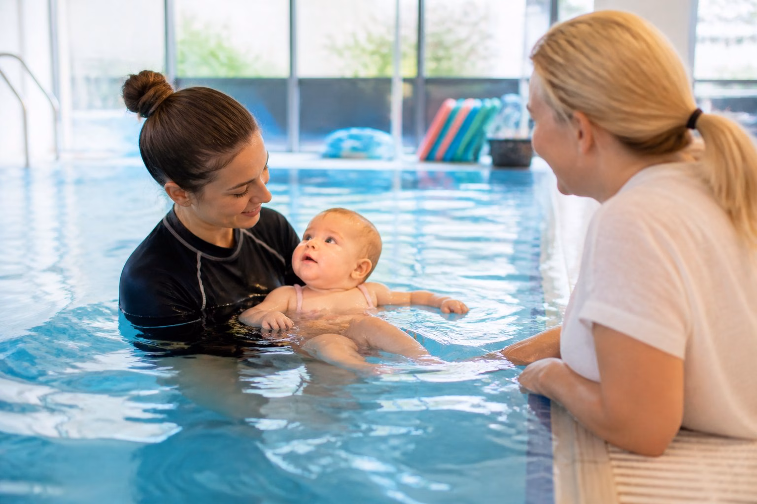 An instructor safely supports a baby during a supervised swimming lesson while a parent watches nearby.