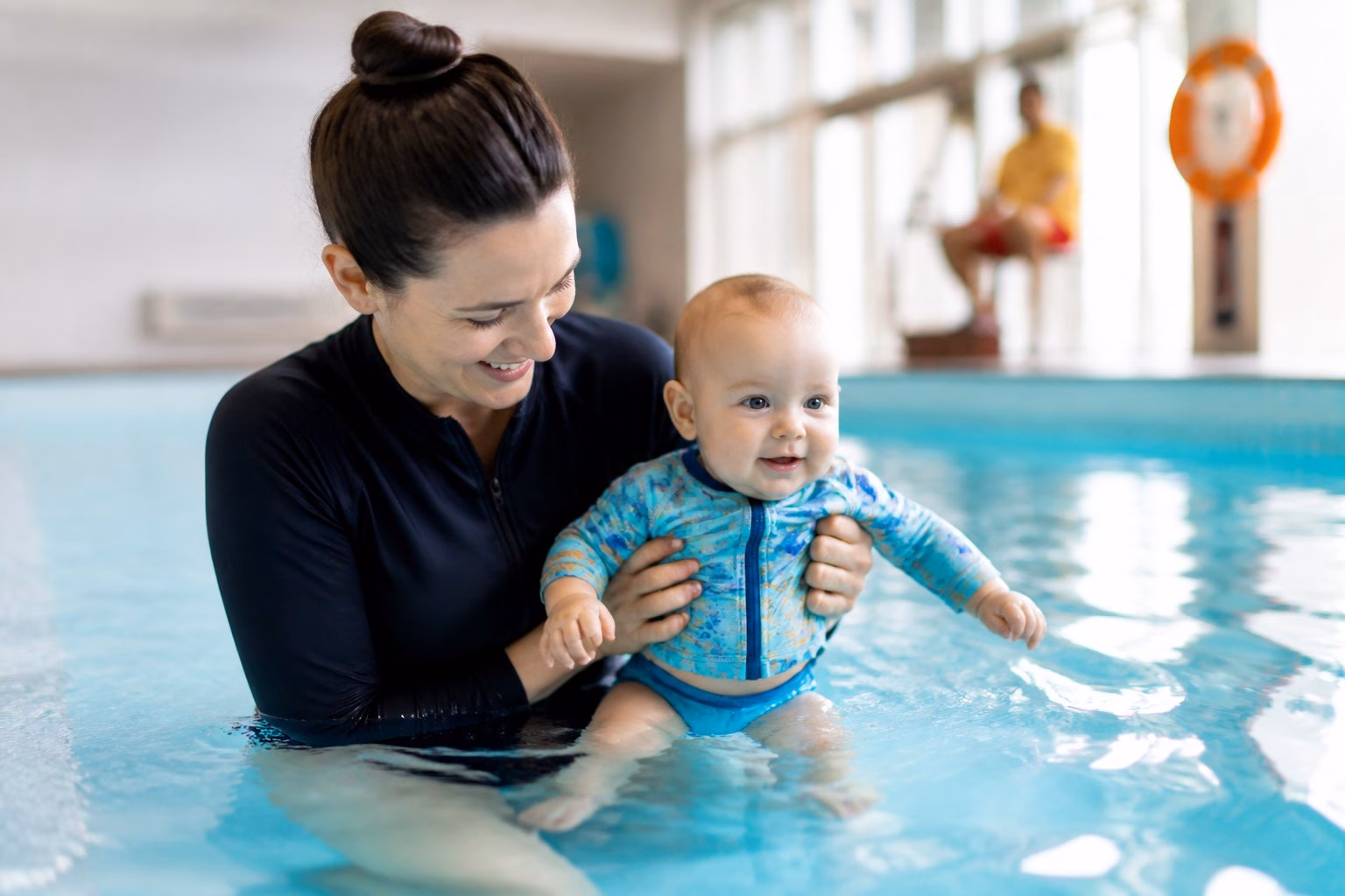 A caregiver closely supervises and supports a baby during a water exercise in a shallow swimming pool.