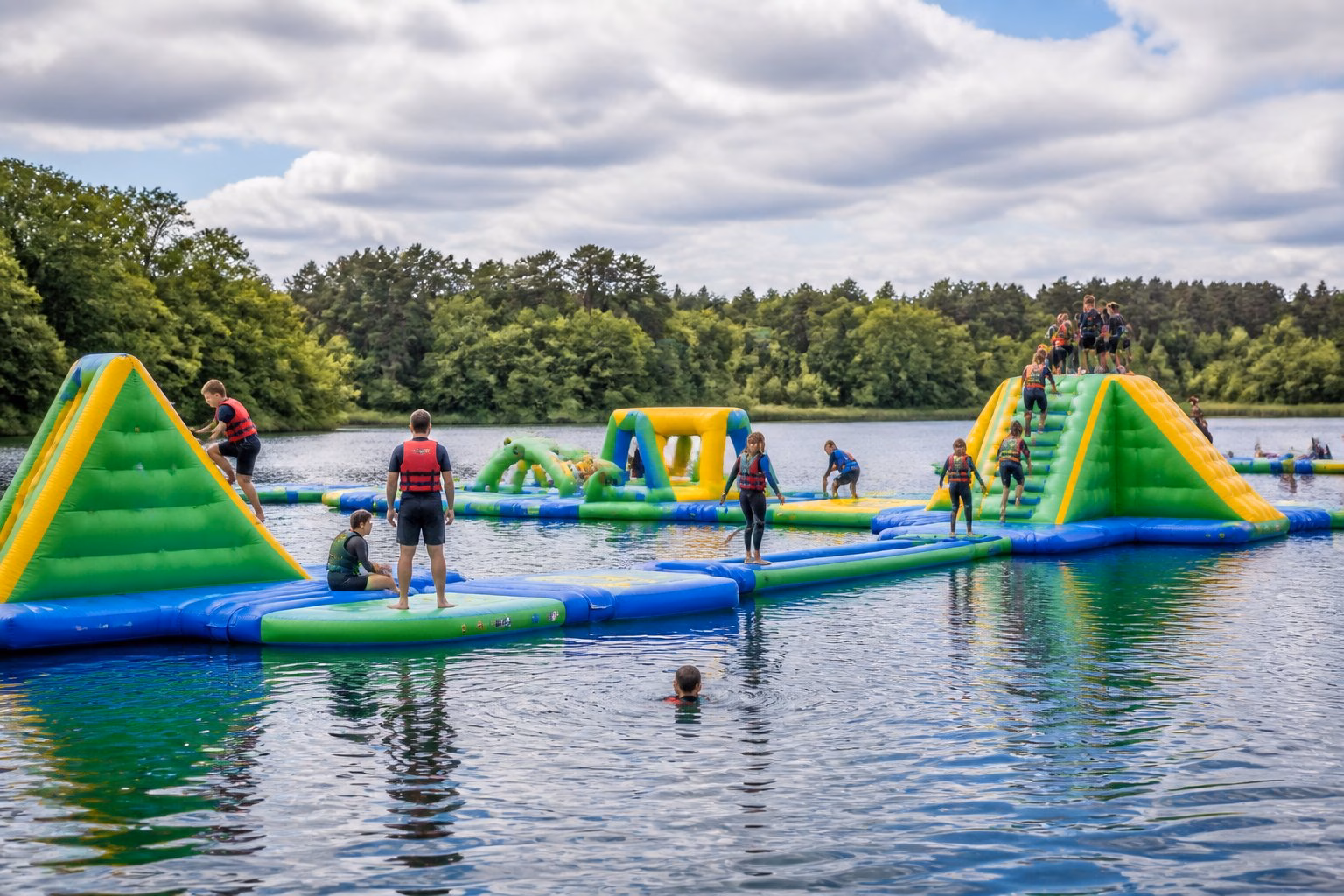 People enjoy a floating inflatable aqua park on a lake surrounded by woodland.