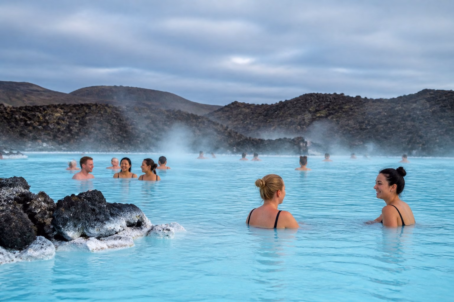 Visitors swim and relax in the steaming turquoise waters of Iceland's Blue Lagoon, surrounded by dark volcanic rocks.