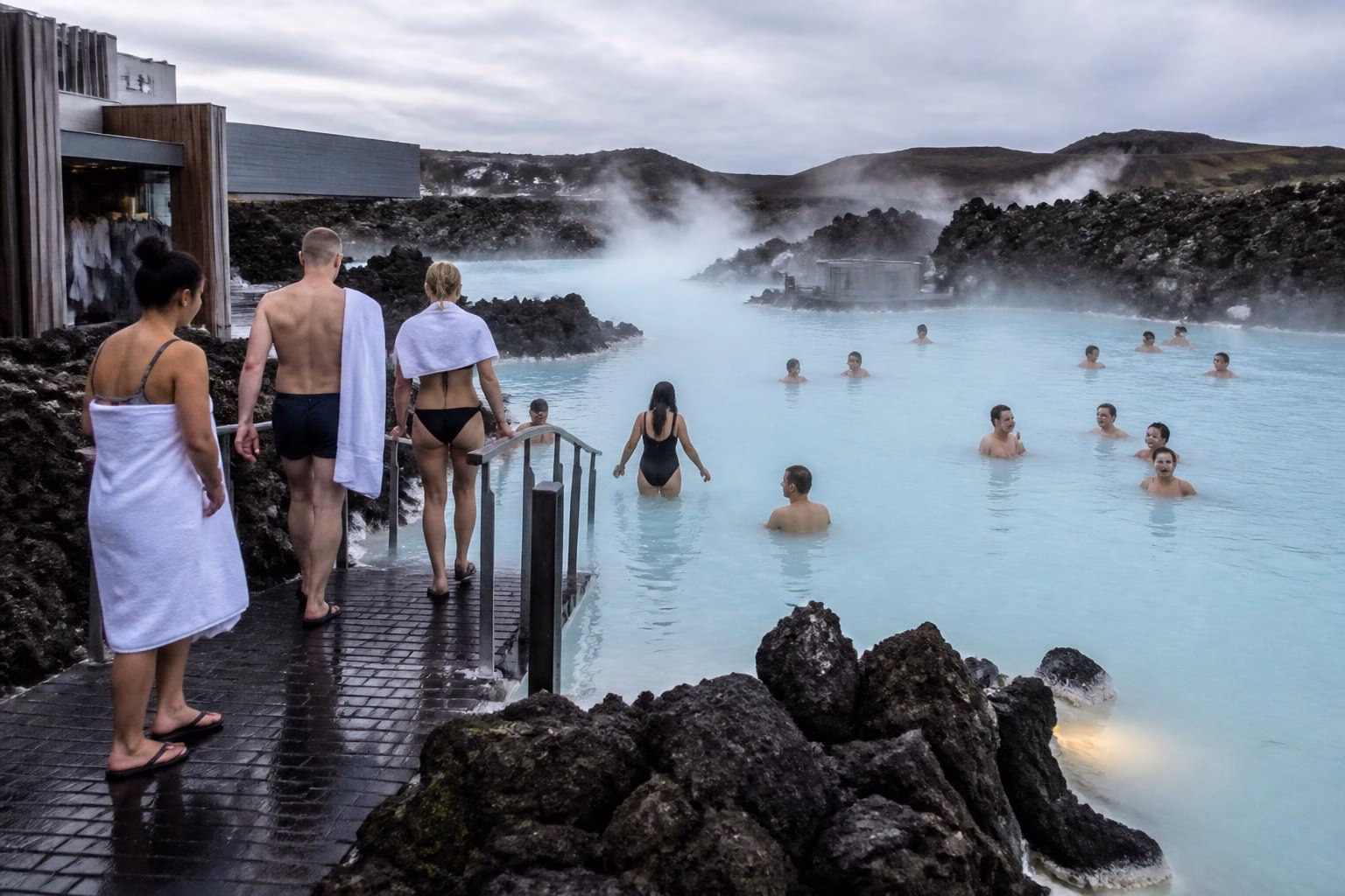 Visitors approach and relax in Iceland’s steaming blue lagoon surrounded by dark volcanic rocks.