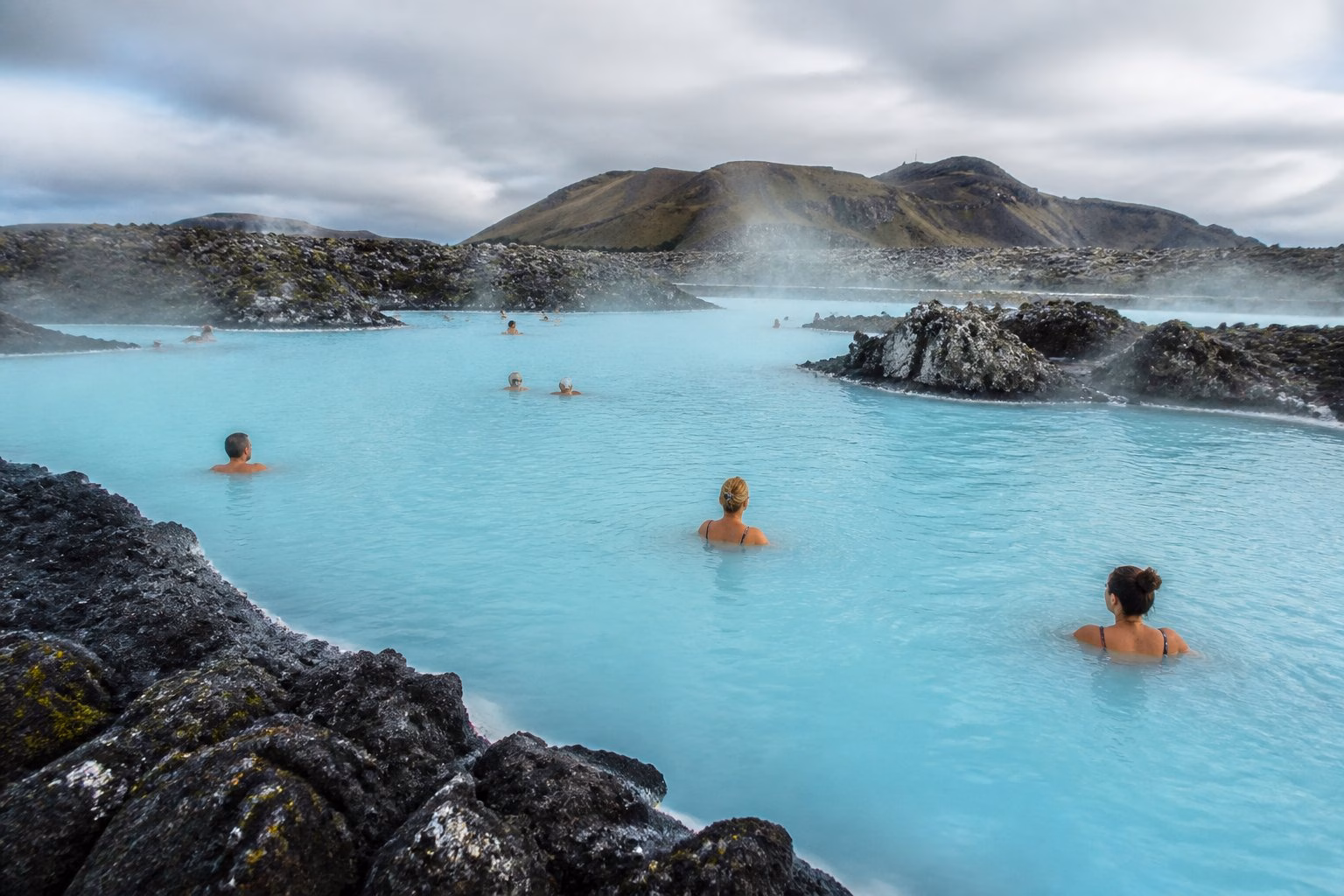 Swimmers relax in the steaming turquoise waters of Iceland’s Blue Lagoon, surrounded by dark volcanic rocks.