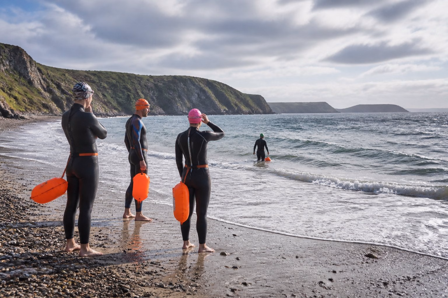 Open-water swimmers prepare to enter choppy sea beside a rugged UK coastline.