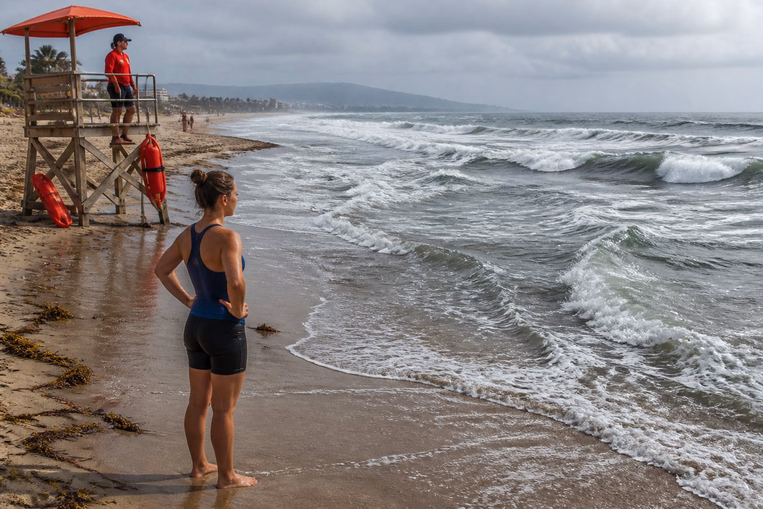 A swimmer pauses on the beach while observing choppy, unsettled waves near a lifeguard station.