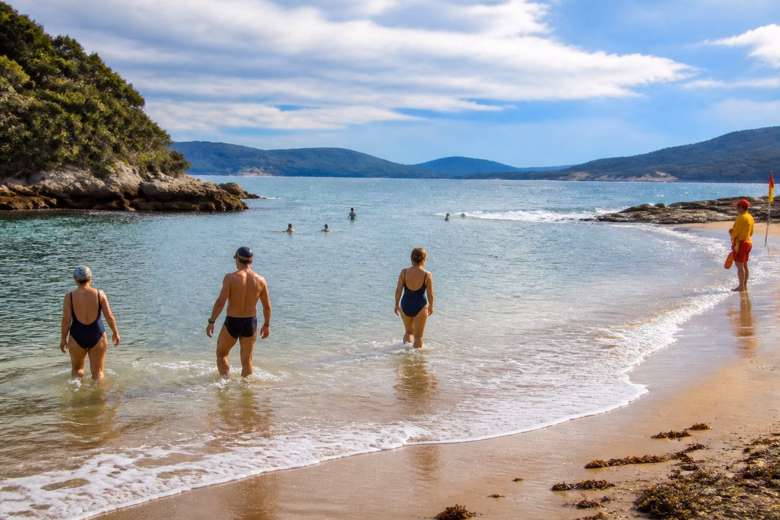 Swimmers enter calm sea water beside a sandy beach with rocky coastal features and changing local conditions.
