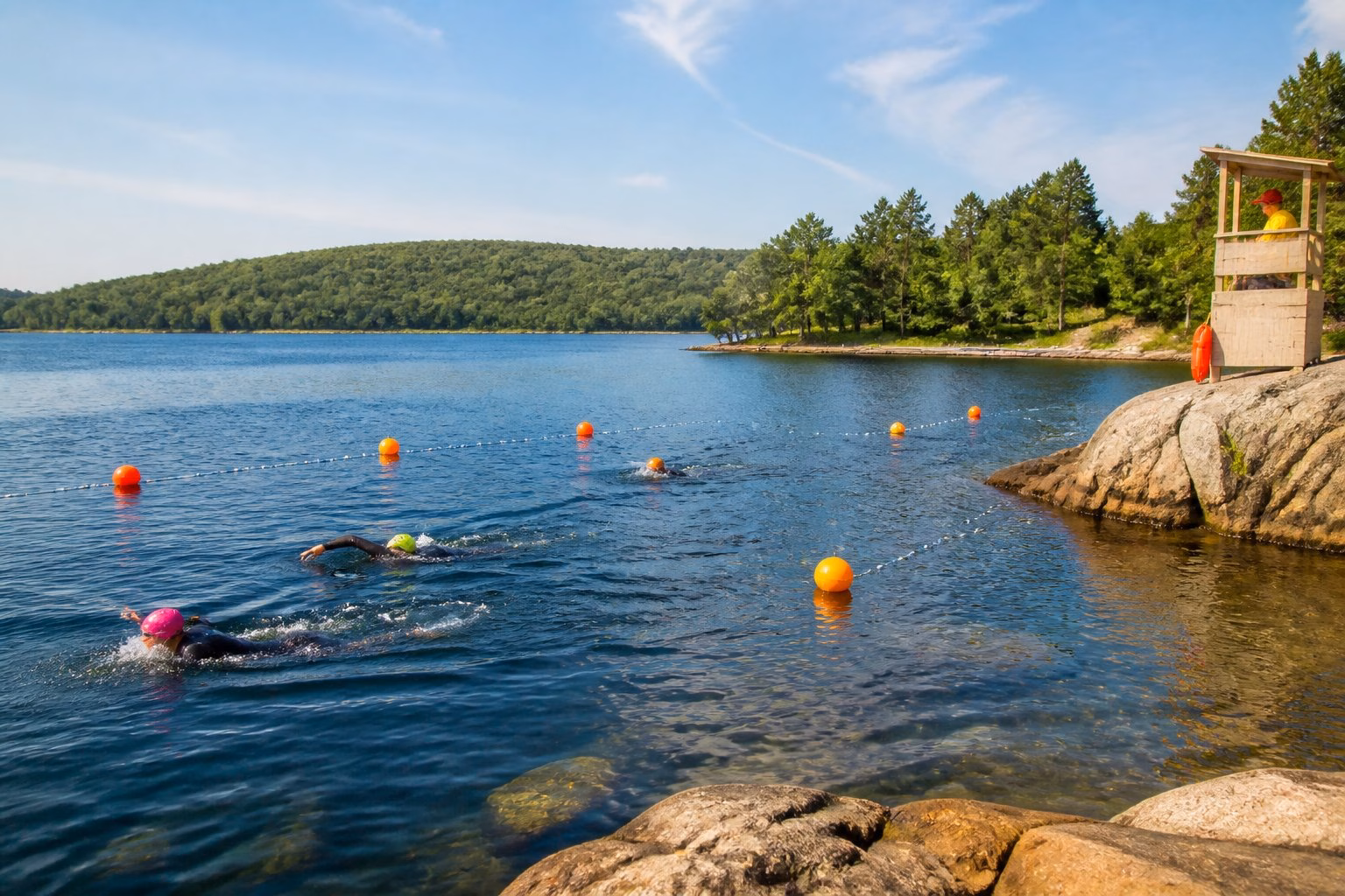 Swimmers in wetsuits enjoying a calm, buoy-marked open-water swimming area at a sunny lake.