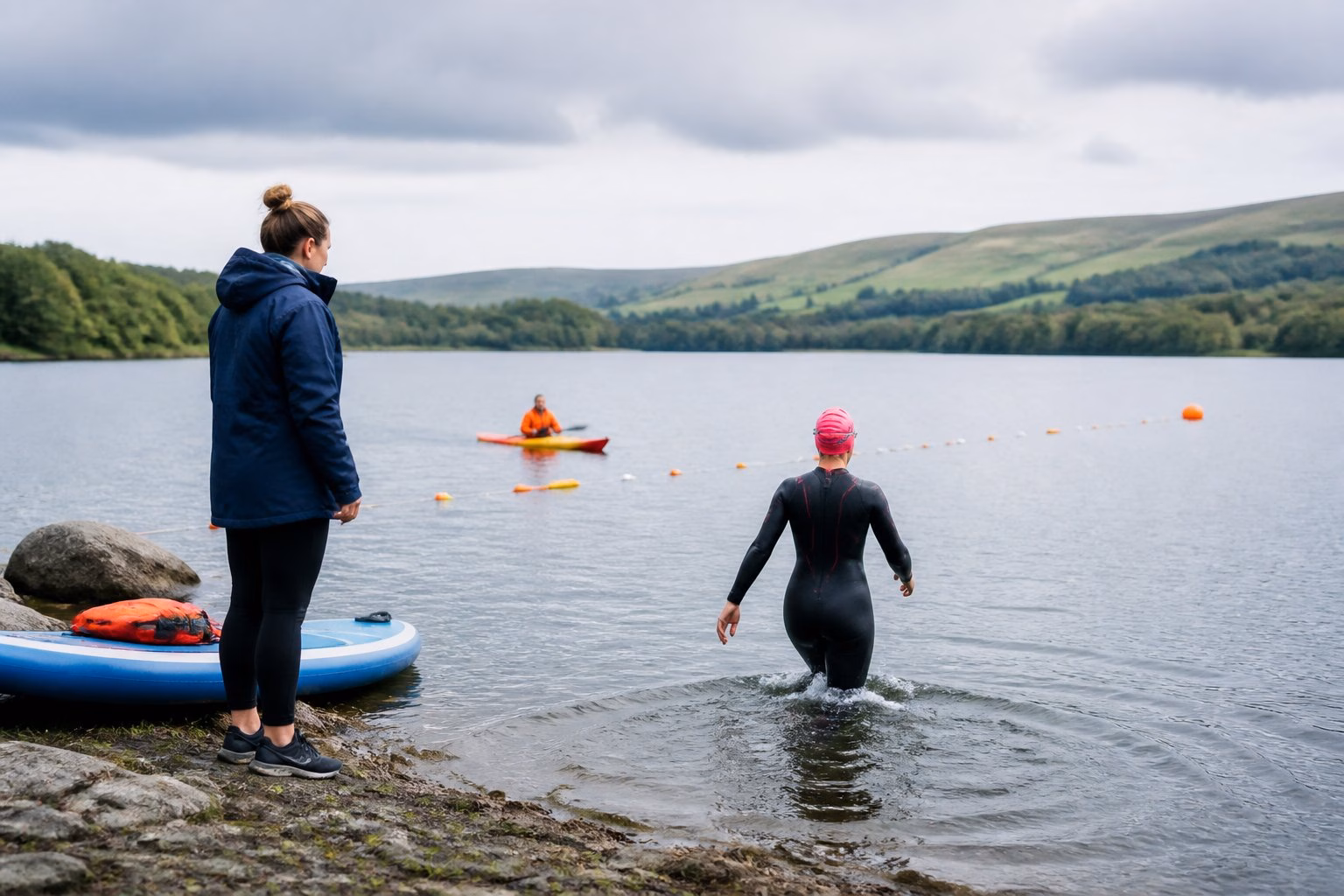 A supervised swimmer enters a scenic open-water lake while a coach waits nearby.