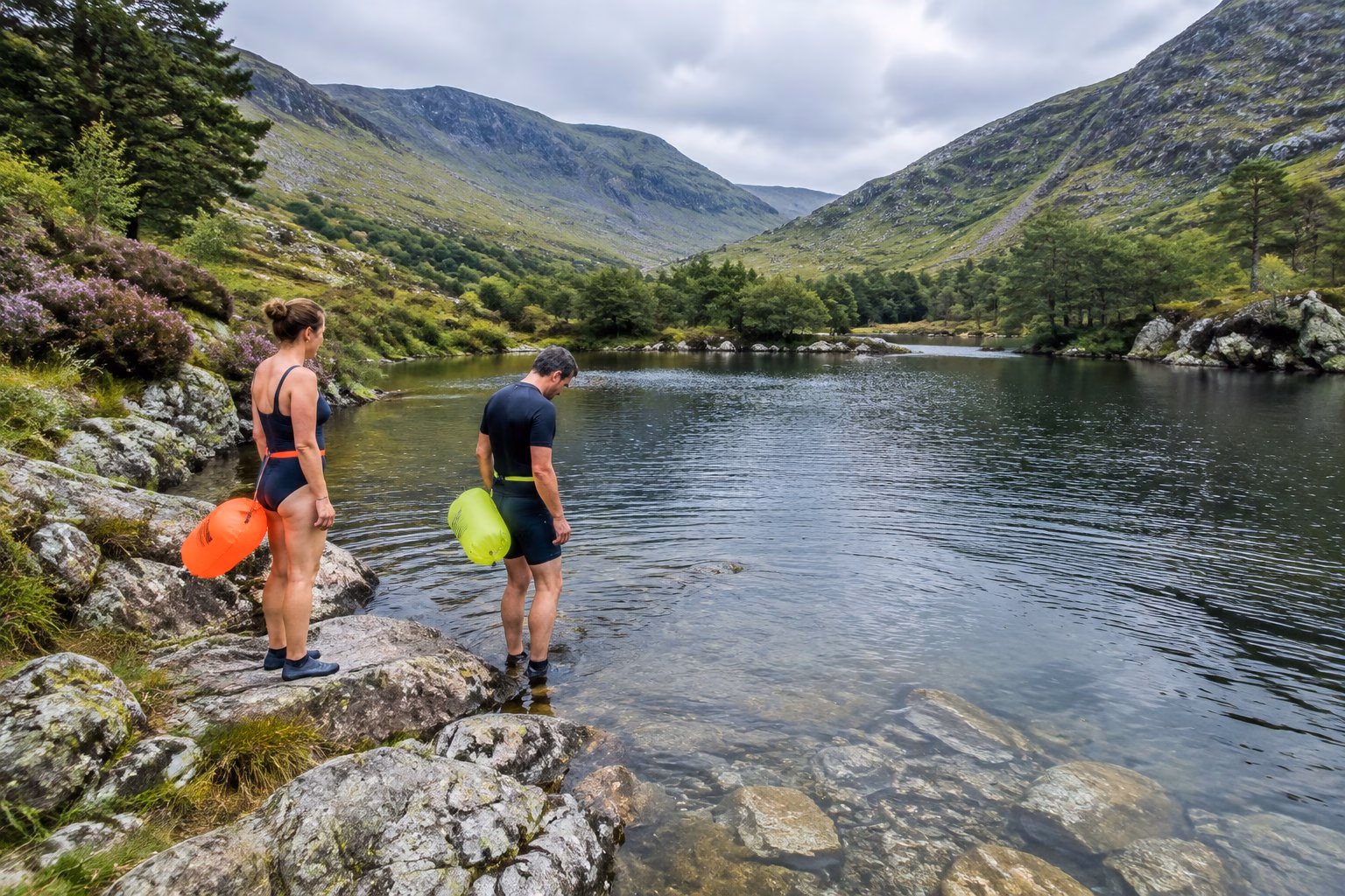 Two swimmers assess a calm mountain pool before entering, surrounded by the green landscape of North Wales.