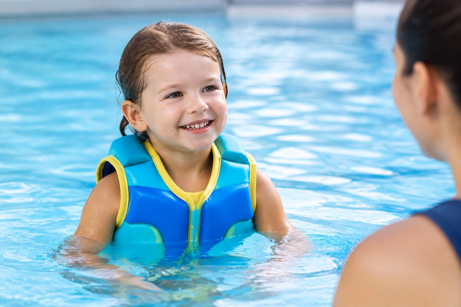 A child wearing a colourful swim vest in a pool with an adult nearby.